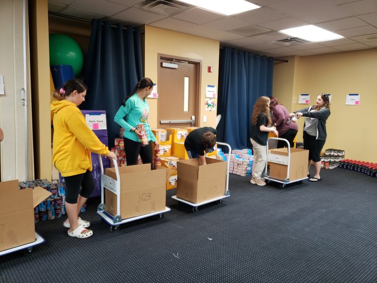 Volunteers pushing carts of boxes past food supplies in a meeting room
