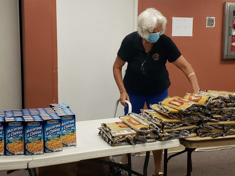 Masked volunteer stacking spaghetti packs beside macaroni and cheese boxes at Monthly Bag Packings