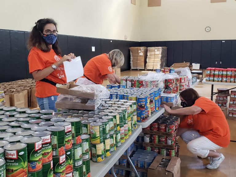 Masked volunteers in orange shirts sorting heaps of canned vegetables at Monthly Bag Packings