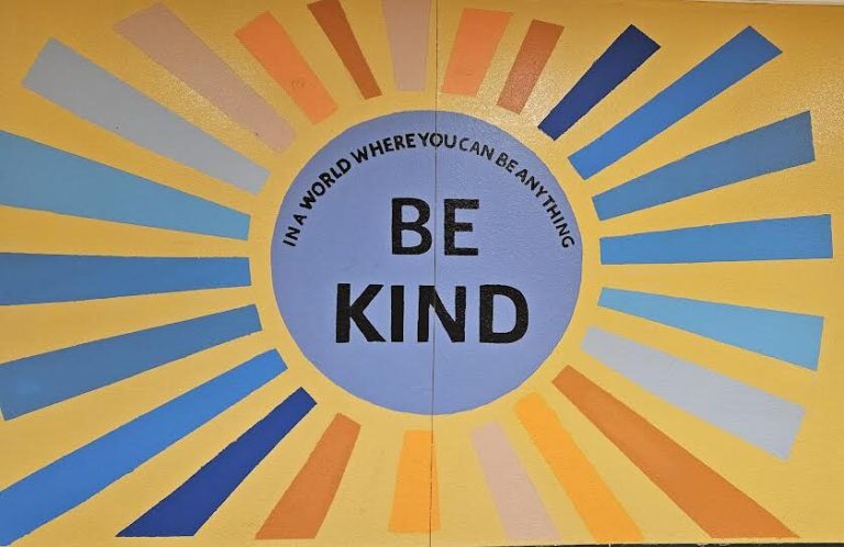 Painted sunburst mural reading In a World Where You Can Be Anything, Be Kind at a Monthly Packings site