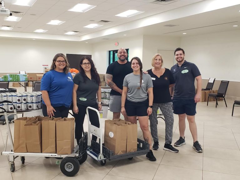 Six volunteers with carts of packed grocery bags in a large hall