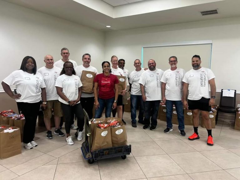 Group of volunteers in white shirts posing behind a cart of filled grocery bags at Monthly Packings