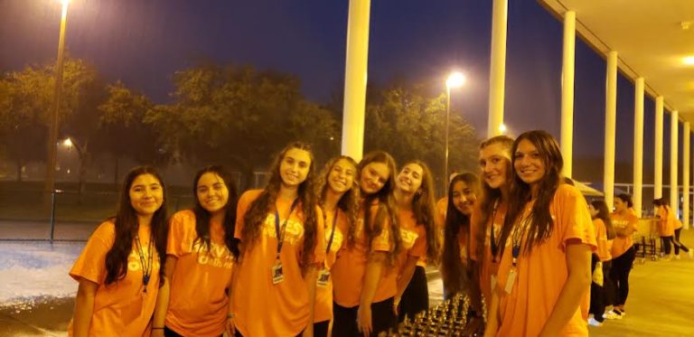 Cypress Bay High School students in orange shirts lined up under a covered walkway at night