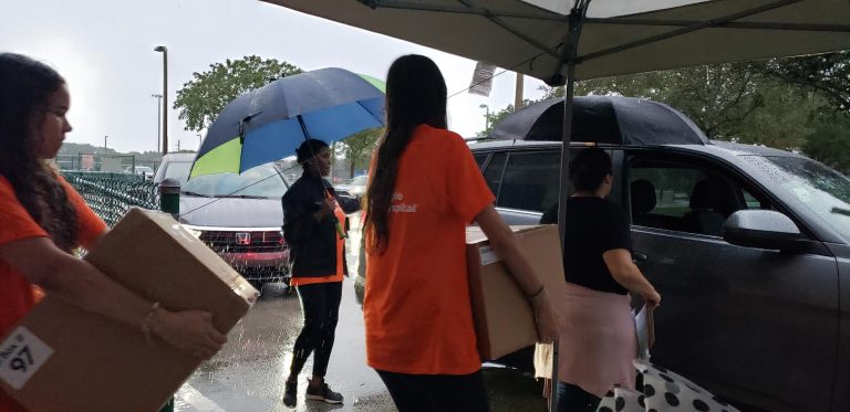 Cypress Bay High School volunteers in orange carrying boxes of food to cars in the rain