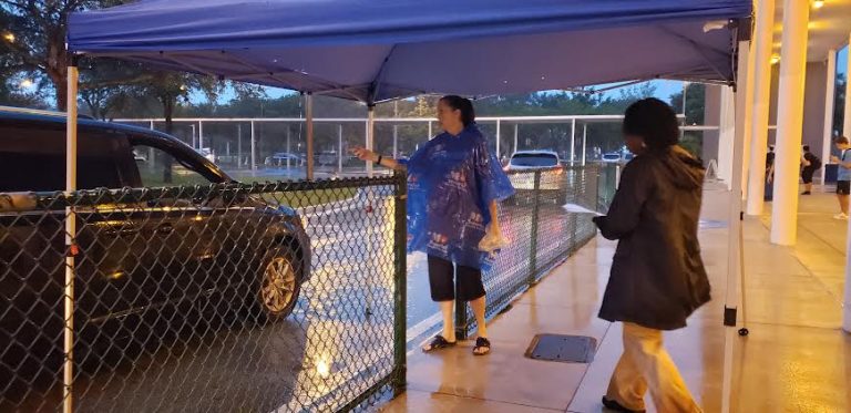 Cypress Bay High School volunteer in a rain poncho greeting a car at a drive-through donation tent