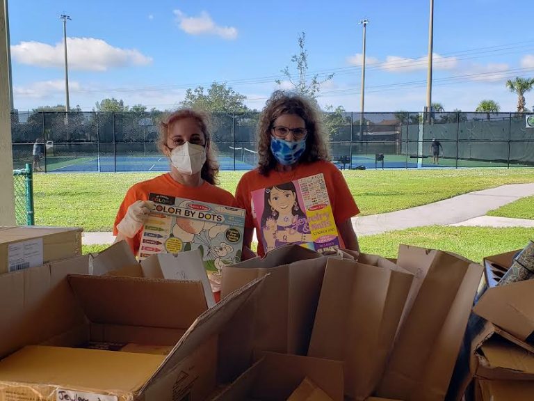 Two masked volunteers holding up coloring books outdoors beside boxes and bags at Monthly Bag Packings