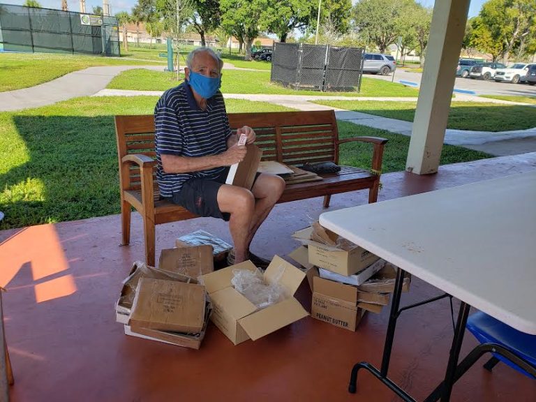 Masked volunteer seated on a bench beside open boxes on a covered patio at Monthly Bag Packings