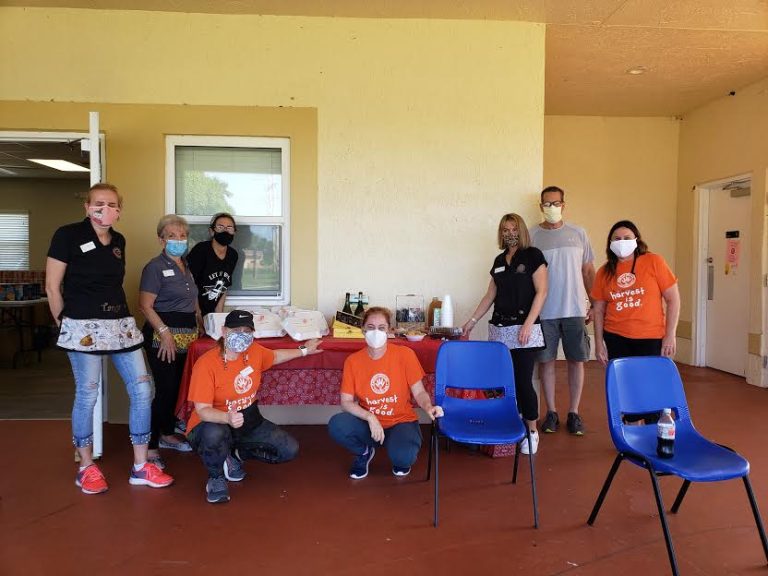 Masked volunteers posing by a table and blue chairs on a covered patio at Monthly Bag Packings