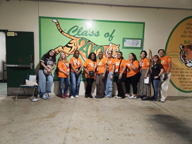 Blanche Ely High School volunteers in orange Harvest Drive shirts posing in front of a tiger class mural