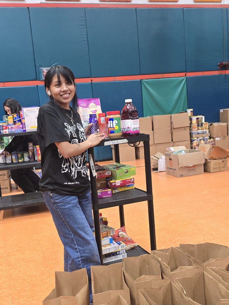 Blanche Ely High School student holding food items beside a cart, with paper bags lined up in front