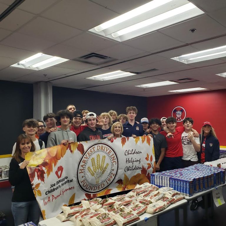 Teens and adults gathered with a Harvest Drive banner behind tables of packaged food