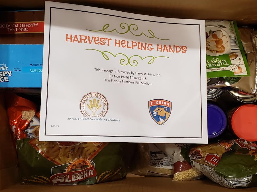 Box of pasta and canned food with a Harvest Helping Hands sign naming the Florida Panthers Foundation