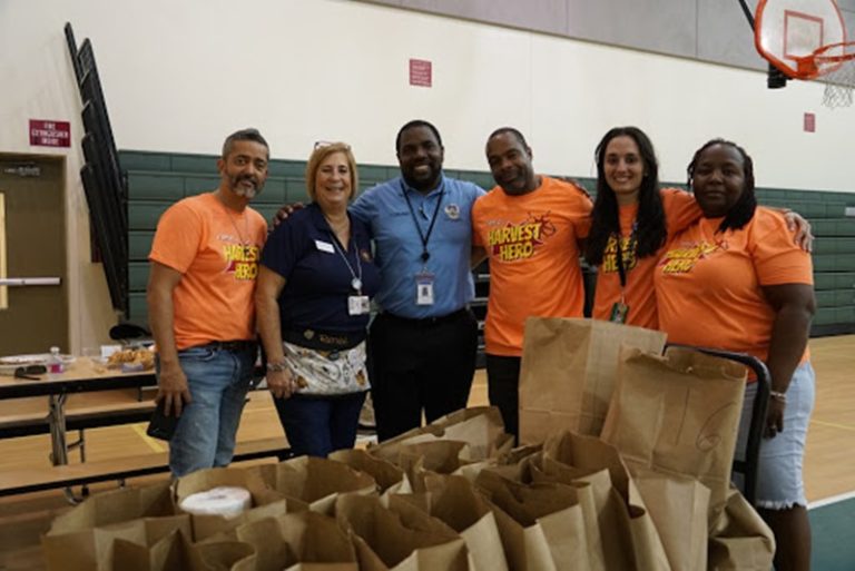 Glades Middle School volunteers in "Harvest Hero" shirts behind a table of paper bags in the gym