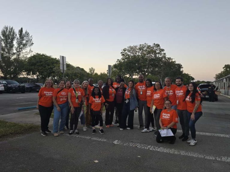 Large group of volunteers in orange shirts posing in the Glades Middle School parking lot at sunrise