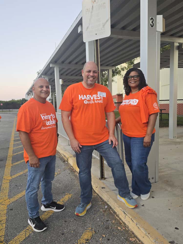 Three volunteers in orange Harvest Drive shirts standing under a covered walkway at Glades Middle School