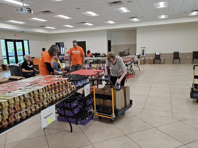 Volunteers in orange shirts filling grocery bags from tables of canned goods during Monthly Packings