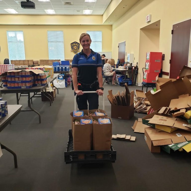 Police officer pushing a cart of grocery bags past tables of food and stacked boxes