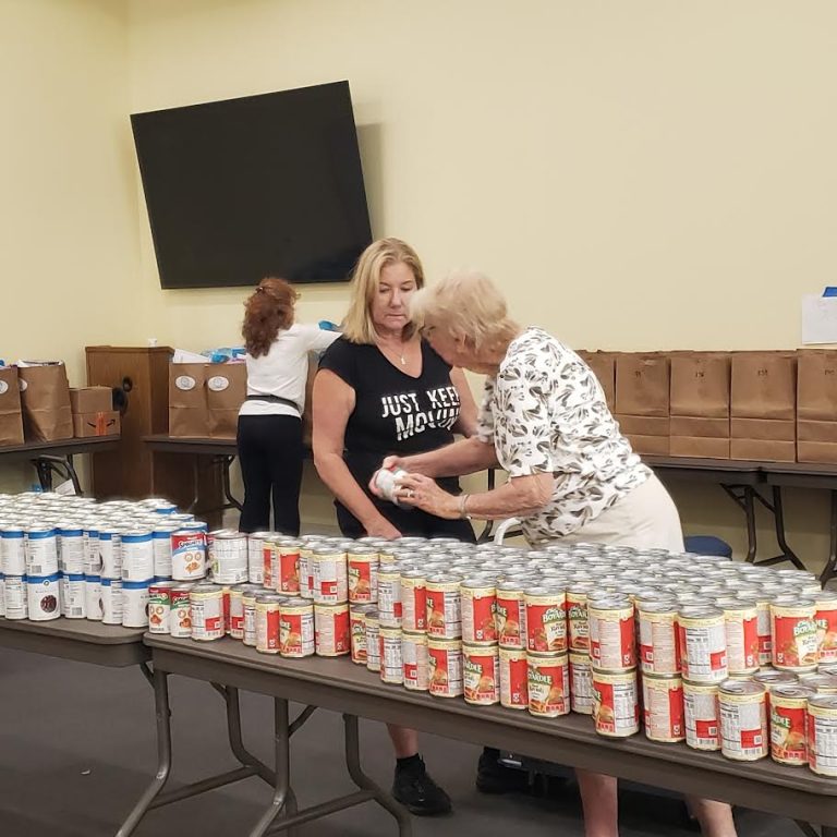 Volunteers sorting canned goods on a long table in front of rows of paper bags