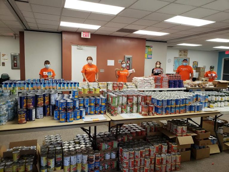 Volunteers in orange shirts behind long tables loaded with canned food at Monthly Bag Packings