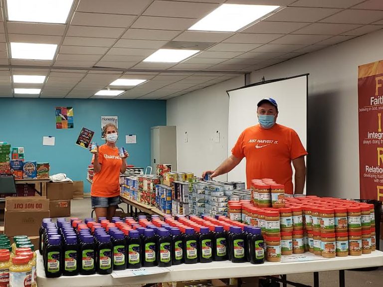 Masked volunteers in orange shirts behind tables of jars and canned goods at Monthly Bag Packings