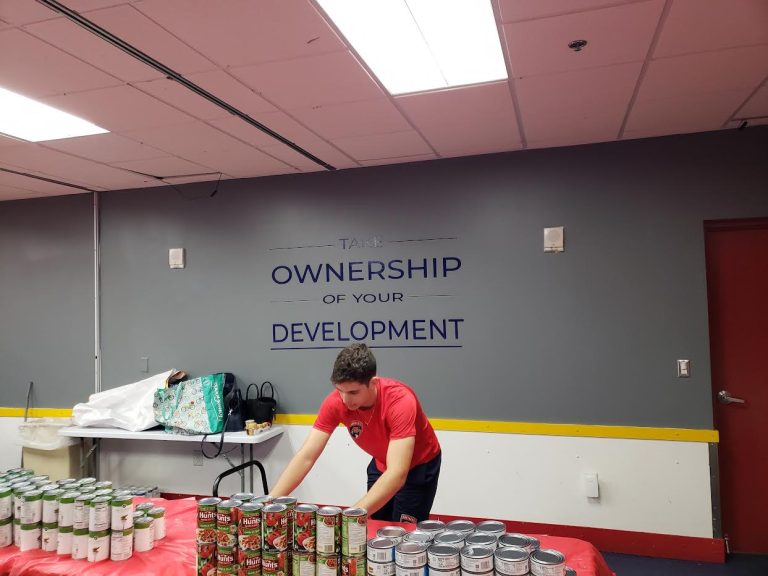 Teen arranging cans on a red-covered table under a Take Ownership of Your Development wall sign