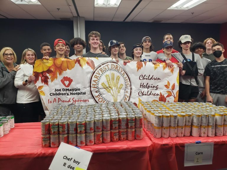 Teens and adults holding a Harvest Drive Children Helping Children banner behind rows of canned goods