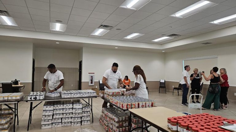 Volunteers working along tables lined with canned food in a large hall at Monthly Packings