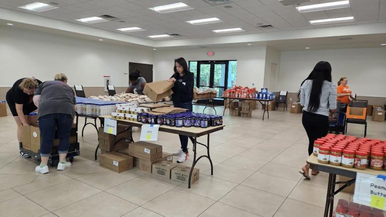 Volunteers bending over boxes at a food table while others restock during Monthly Packings