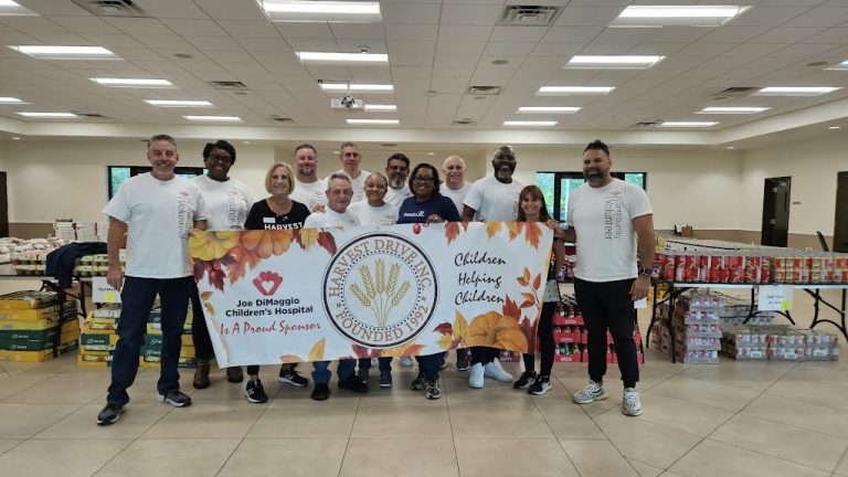 Volunteers in white shirts holding a Harvest Drive banner in a hall of food at Monthly Packings