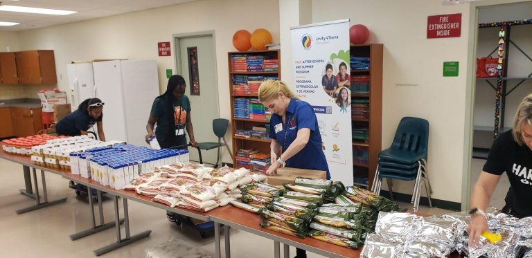 Volunteers and students packing snack bags at classroom tables beside a banner stand at Monthly Packings