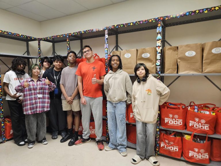Students lined up in a school pantry in front of shelves of red Goals for Food bags