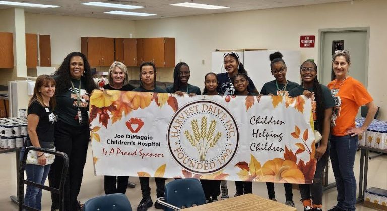 Students and adults holding a Harvest Drive Children Helping Children banner in a classroom at Monthly Packings