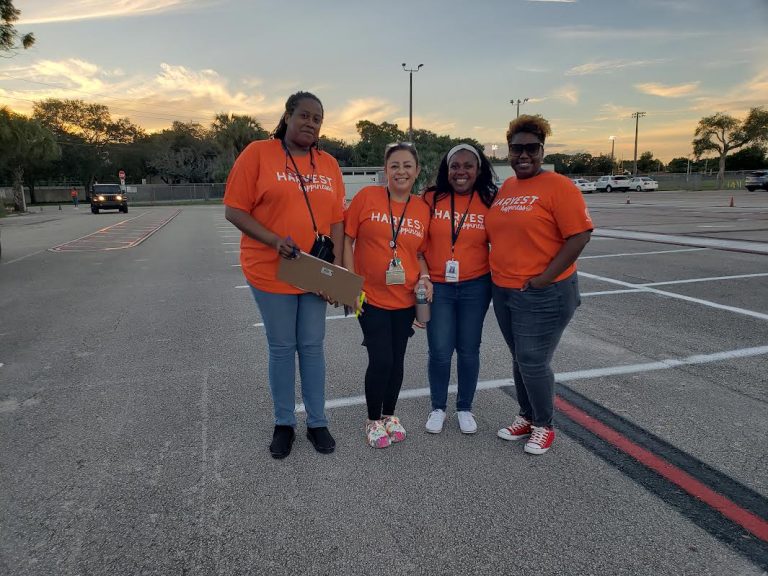 Four Plantation High School volunteers in orange shirts standing together in the parking lot at dusk