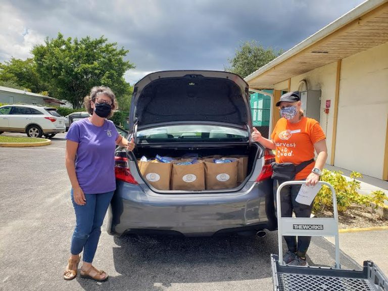 Two masked volunteers beside a car trunk filled with bags and a hand cart at Monthly Bag Packings