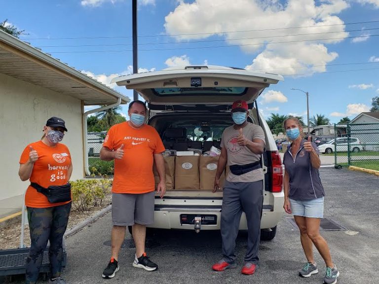 Masked volunteers giving thumbs up behind an SUV loaded with grocery bags at Monthly Bag Packings