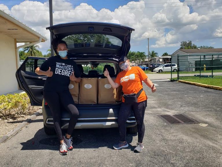 Two masked volunteers seated in an open car trunk holding grocery bags at Monthly Bag Packings