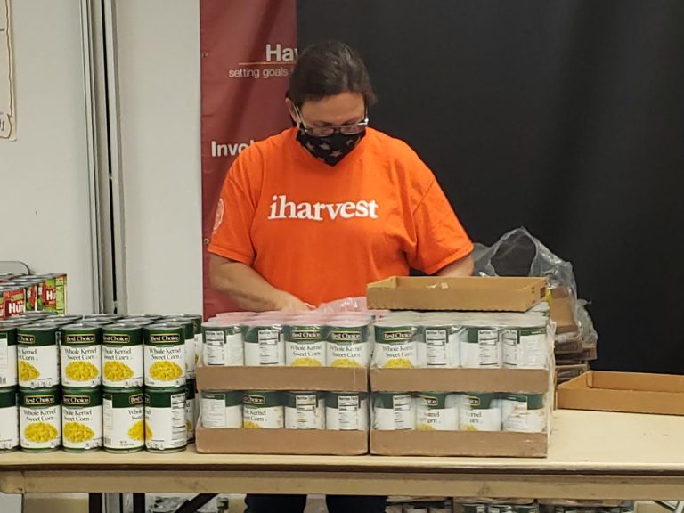 Masked volunteer in an orange shirt unpacking cases of canned corn at Monthly Bag Packings