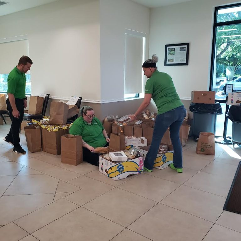 Publix volunteers in green shirts packing brown grocery bags in a community room