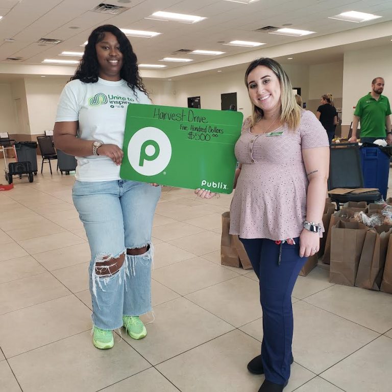 Two women holding a green Publix sign in a hall with grocery bags behind them