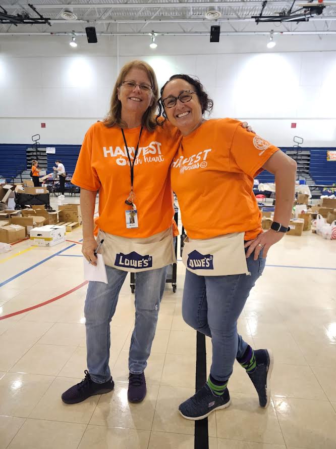 Two Westglades Middle School volunteers in orange Harvest shirts and Lowe's aprons on the gym floor