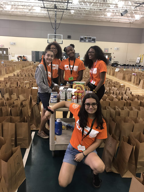 Students in orange Harvest Drive shirts with rows of packed grocery bags in a gym
