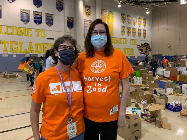 Two Westglades Middle School volunteers in orange "harvest is good" shirts and face masks in the gym