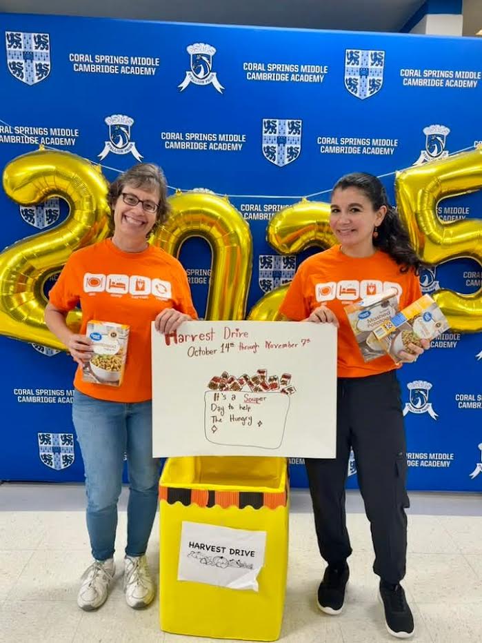 Two volunteers in orange shirts with gold "2025" balloons, a donation poster and a yellow Harvest Drive collection box
