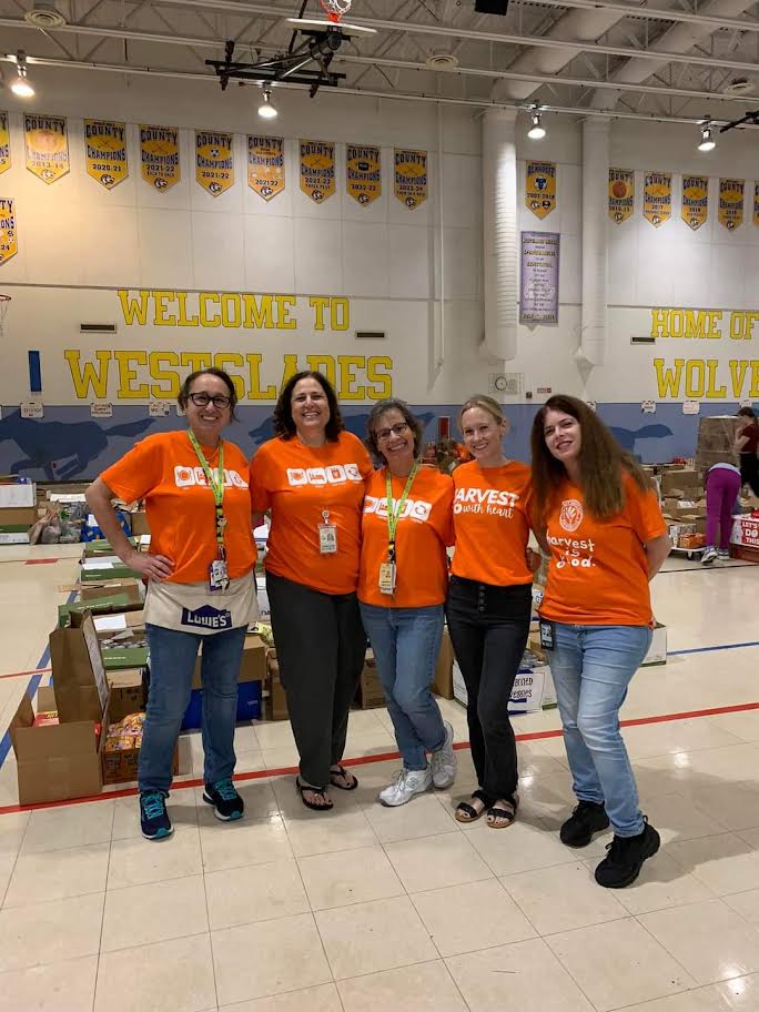 Five Westglades Middle School volunteers in orange shirts standing below the "Welcome to Westglades" sign