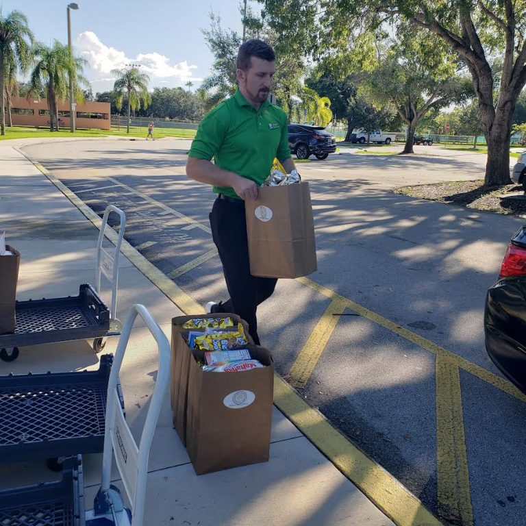 Publix volunteer carrying a grocery bag across a parking lot toward a loaded cart